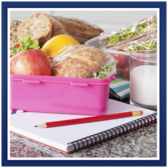 Lunchbox with food inside, a glass of milk and a notebook with a pencil on top of it