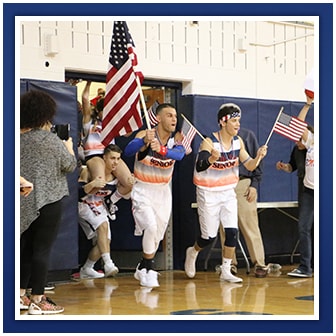 Students wearing patriotic clothes run into a gym holding American flags