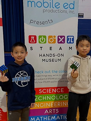 two students holding Rubik's cubes in front of a STEAM Hands-on Museum sign